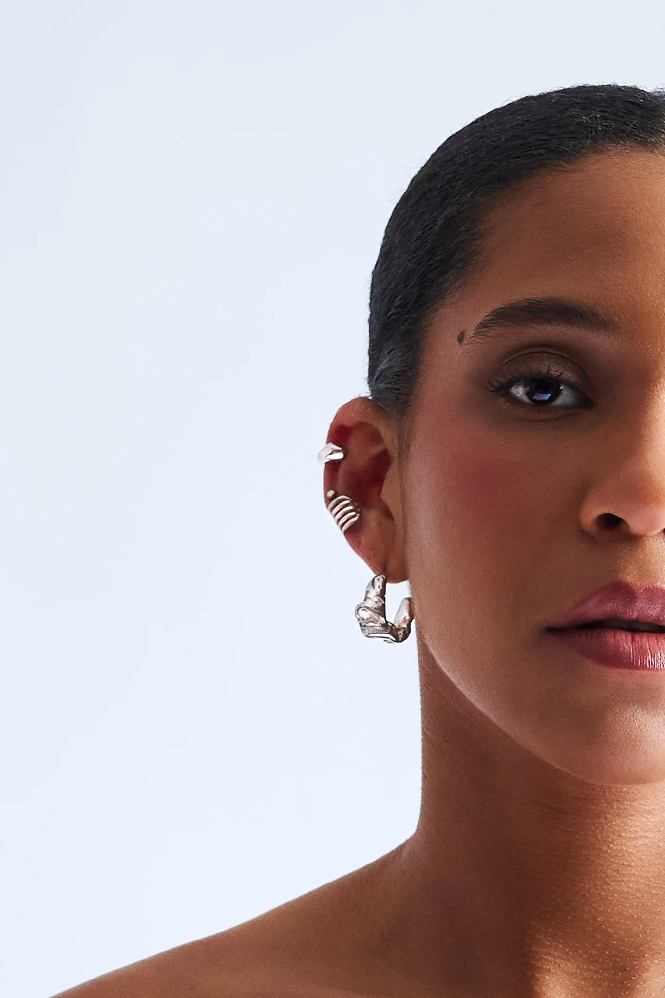 Woman wearing silver sculptural hoop earrings and ear cuffs against light background