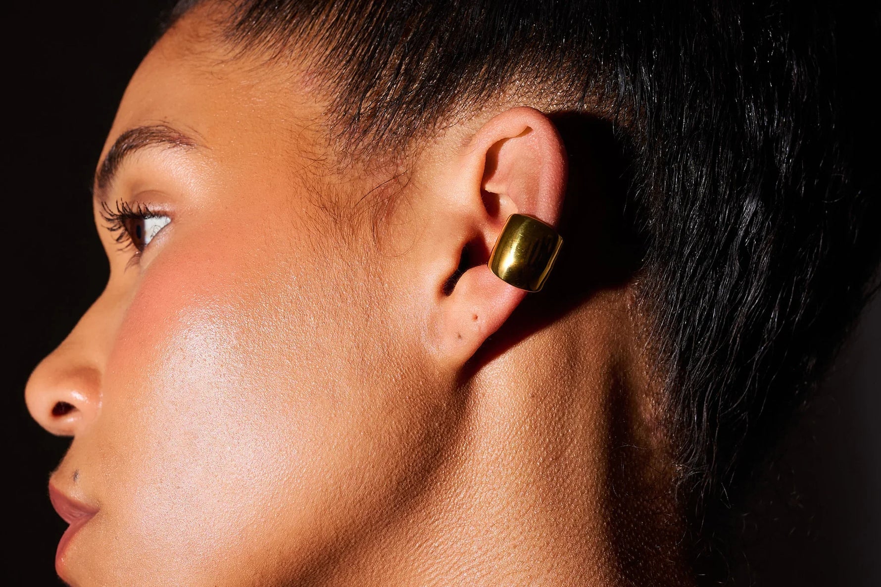 Close-up of a woman wearing a chunky gold ear cuff earring against a dark background