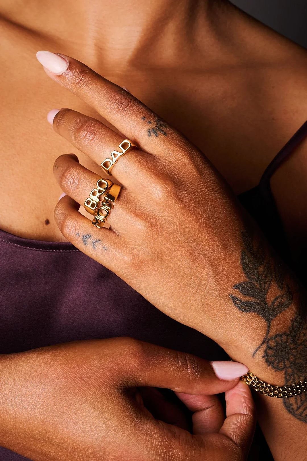 Hand with gold rings spelling BRO, MOM, DAD, floral tattoos, and a gold chain bracelet