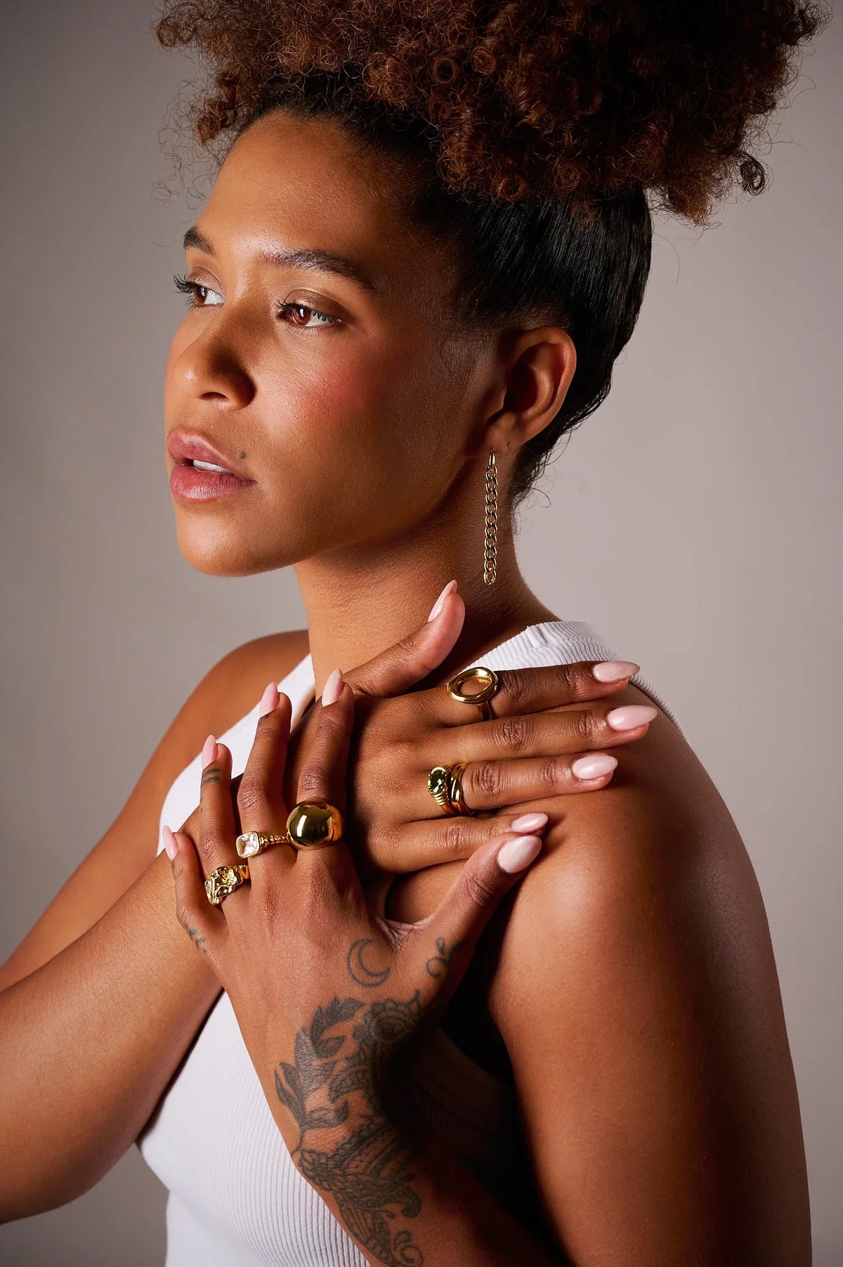 Woman wearing gold rings and a chain earring, with a white top and floral tattoo on hand