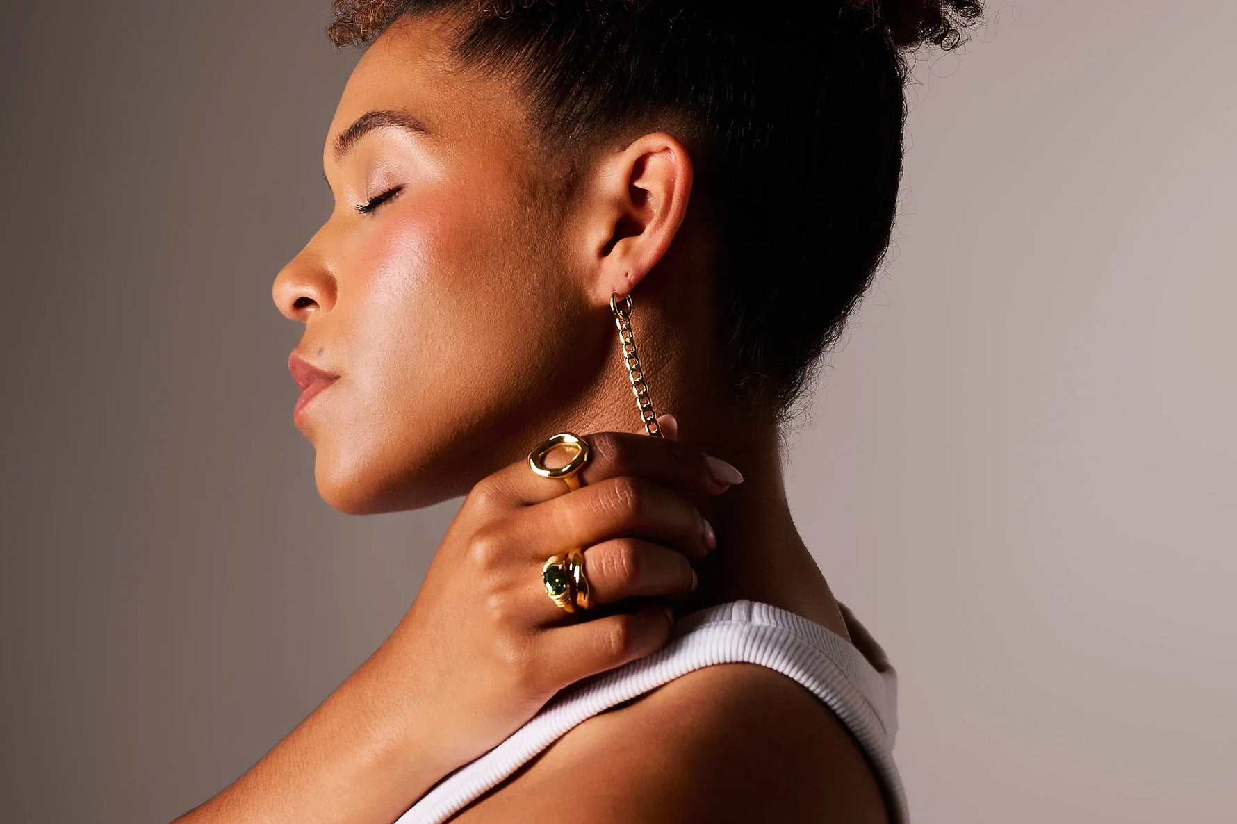 Woman wearing gold chain drop earring and gold cocktail rings, neutral background