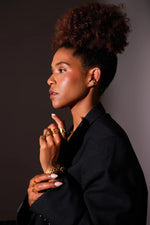 Woman wearing gold jewelry rings, earrings, and bracelet, black blazer, dark background