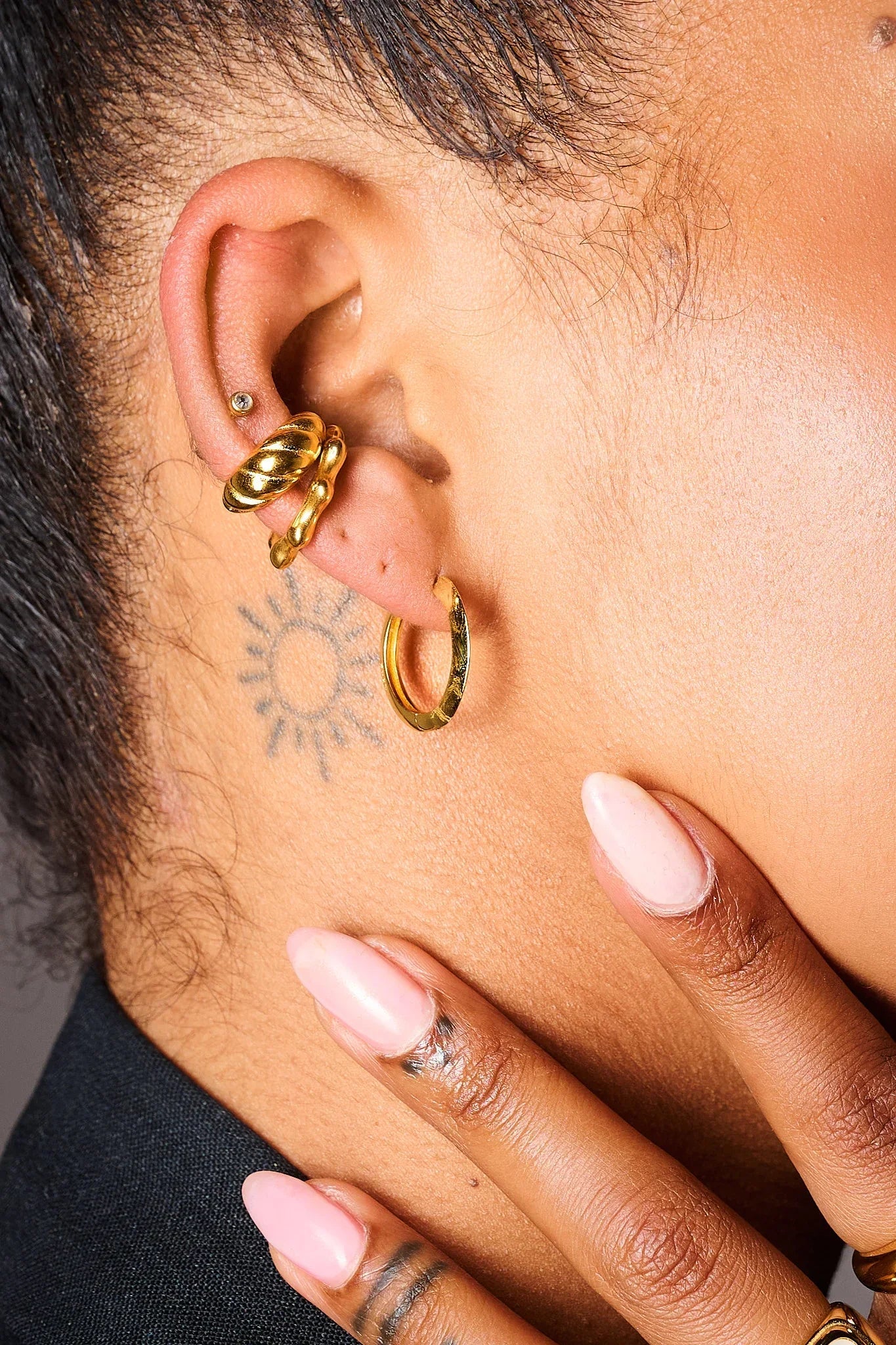 Close-up ear with gold hoop, chunky and textured ear cuffs, diamond stud, sun tattoo, manicured nails