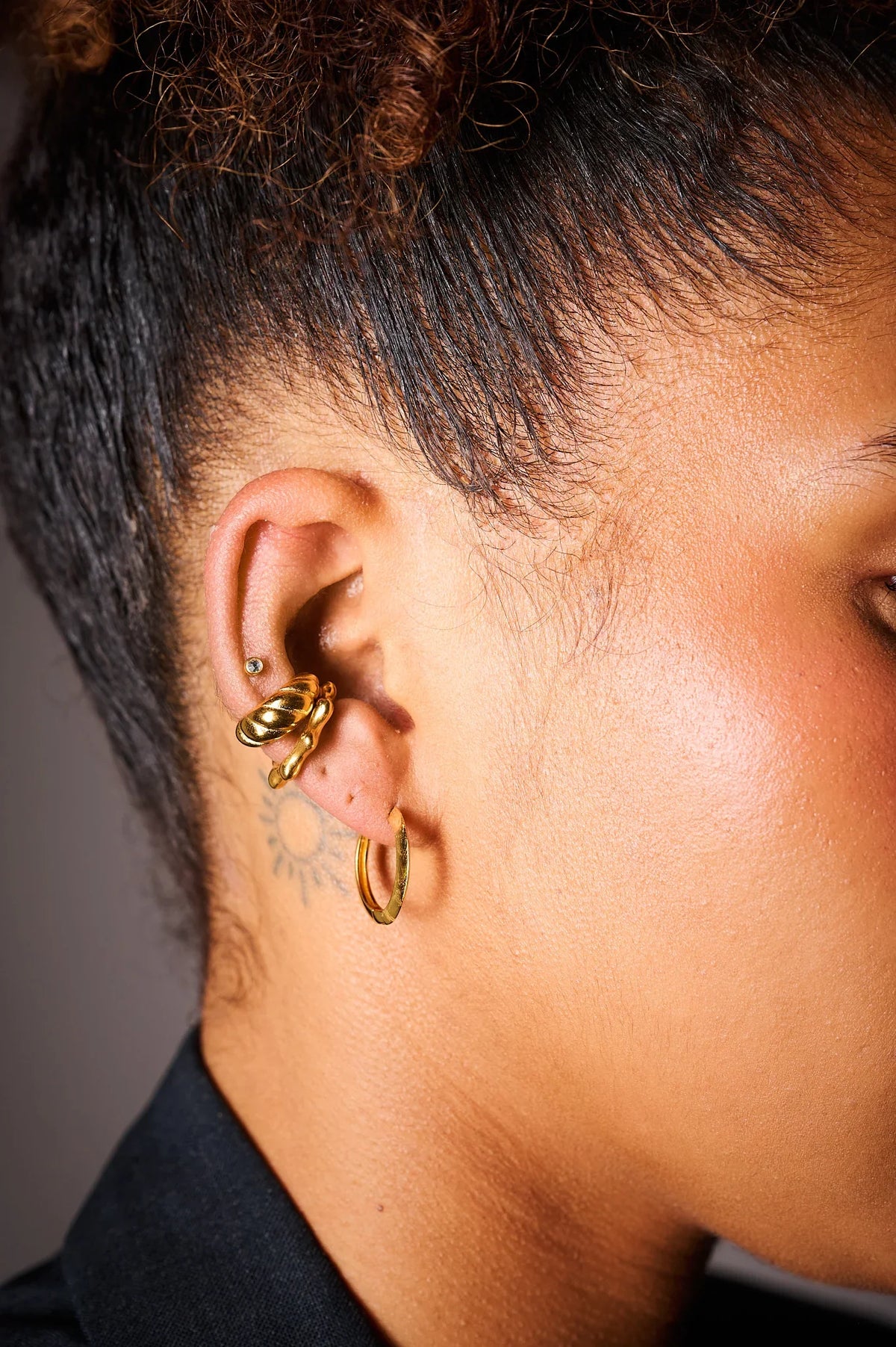 Close-up of ear with multiple gold hoop earrings and a small sun tattoo behind the ear