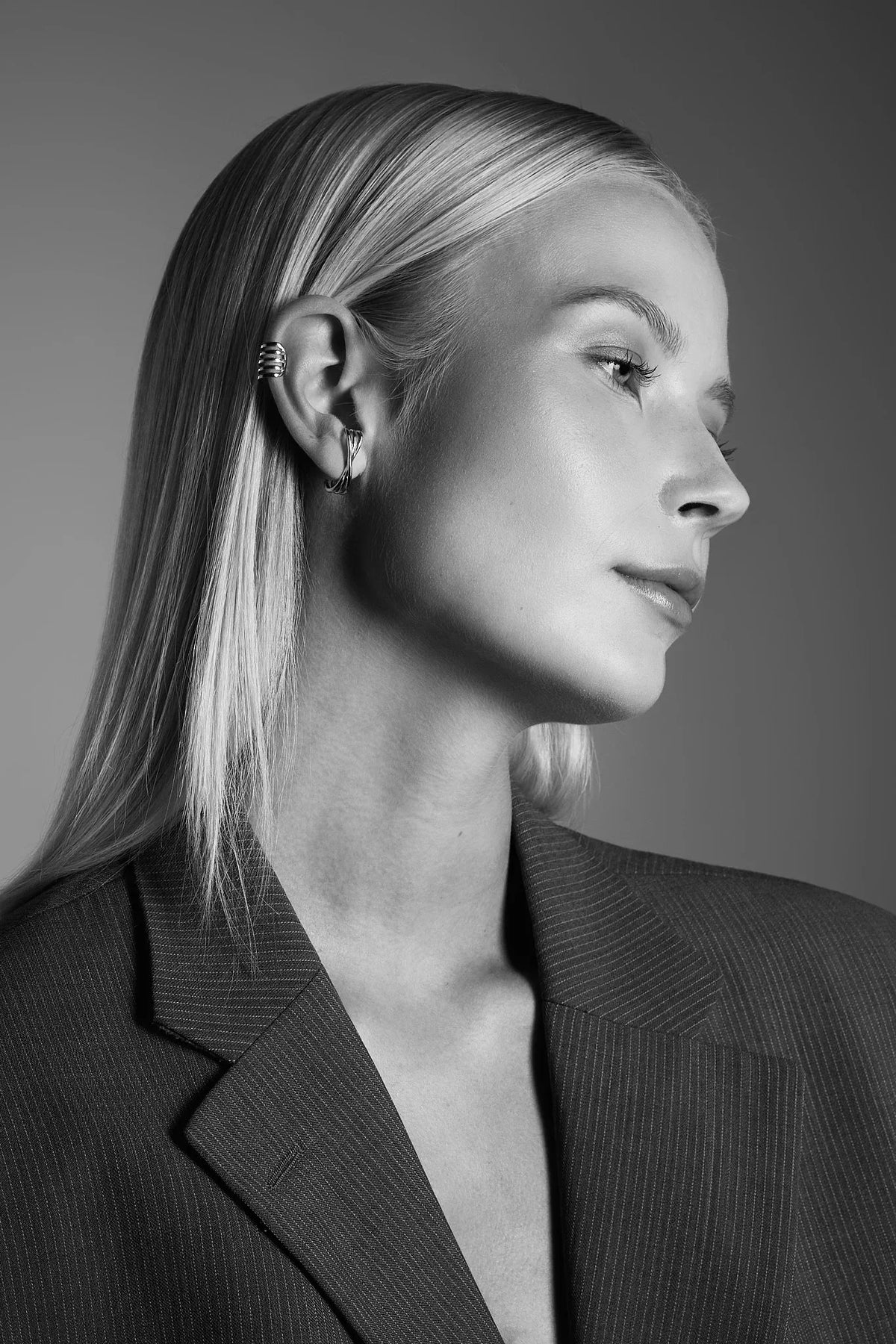 Woman with sleek blonde hair wearing silver ear cuff and hoop earring, dressed in pinstripe blazer