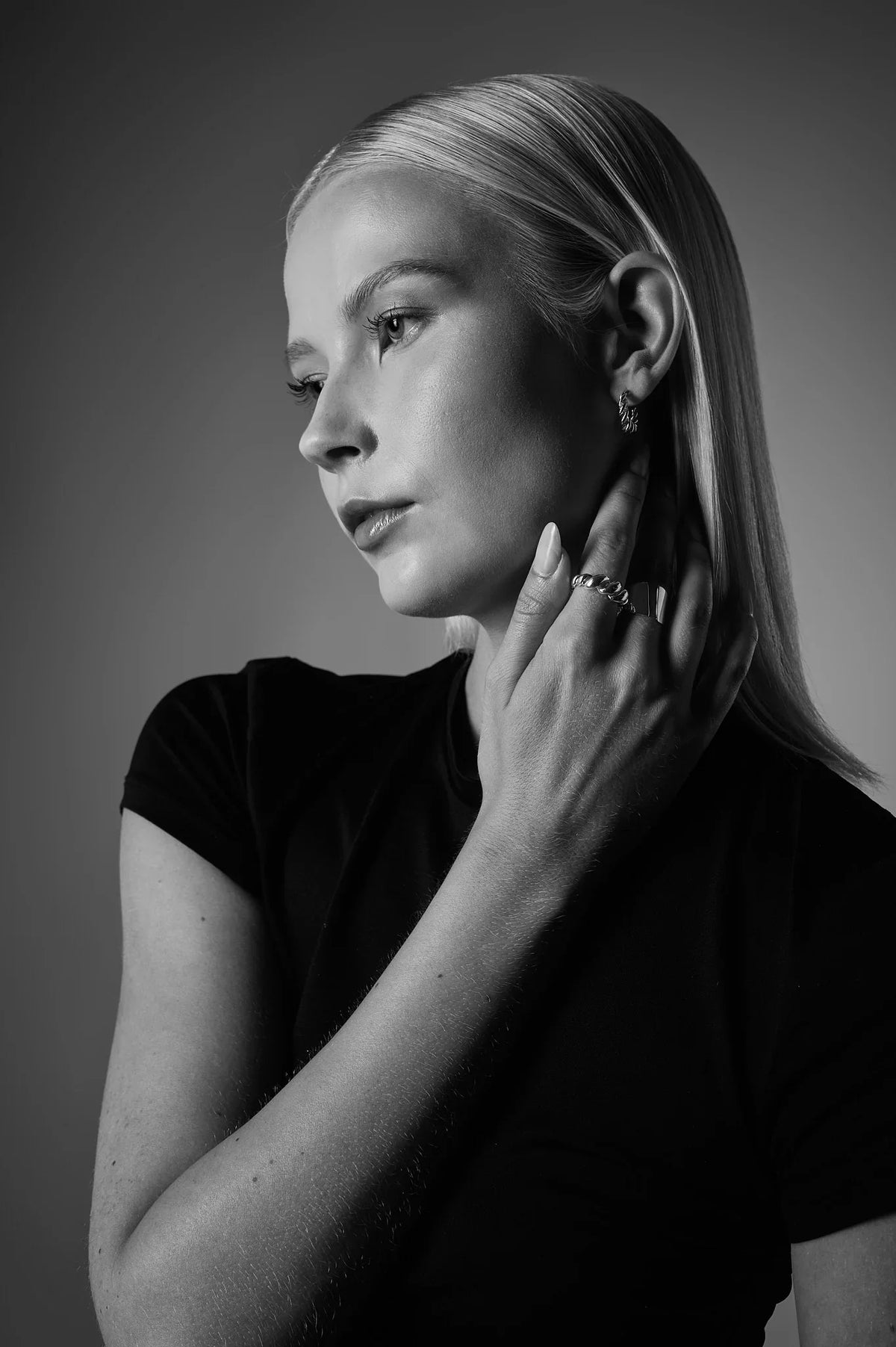 Woman wearing silver ring and hoop earring, hand to neck, black shirt, studio lighting