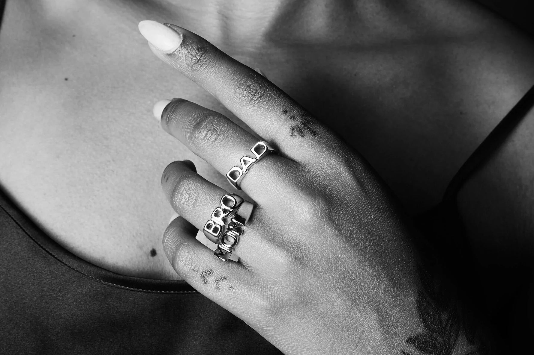 Close-up of hand with silver rings spelling DAD, BRO, MOM, and finger tattoos, on bare chest