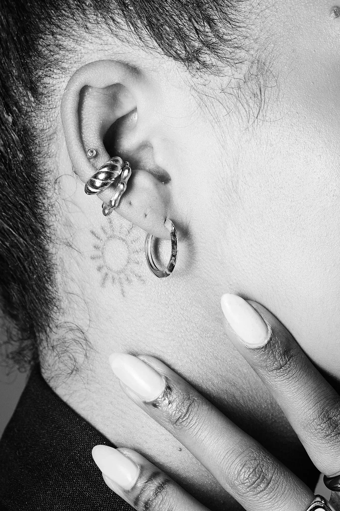 Close-up of ear with multiple silver earrings and sun tattoo, hand with manicured nails
