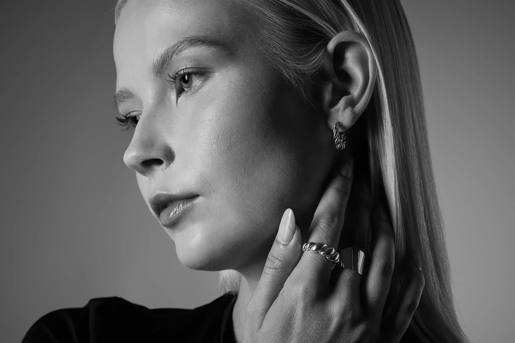 Woman wearing twisted silver hoop earring and chunky silver rings, close-up, black and white