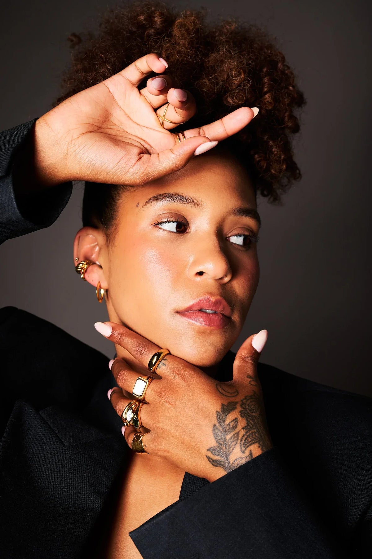 Woman wearing multiple gold rings, gold hoop earrings, and a black blazer, with tattooed hand