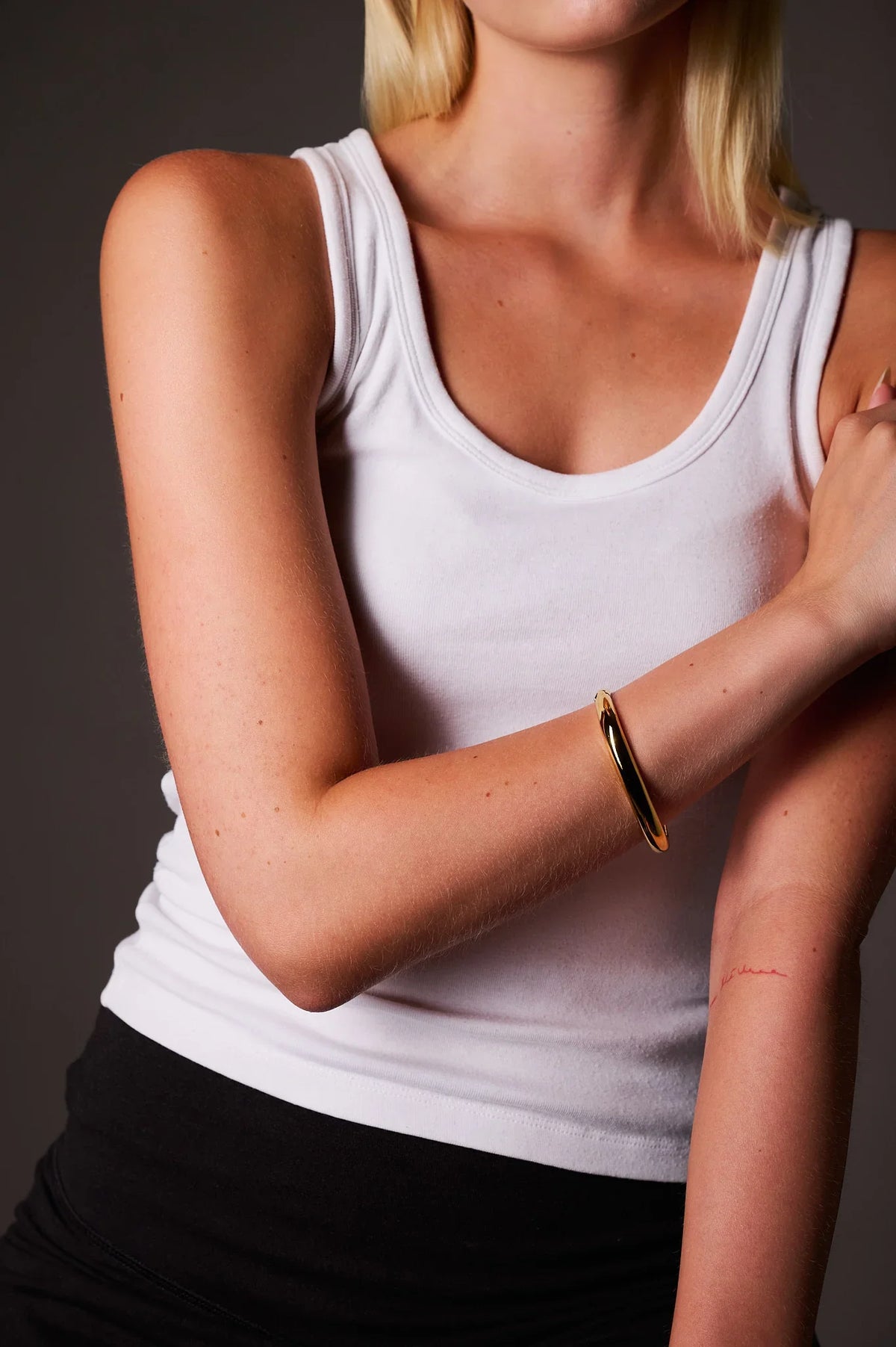 Woman wearing minimalist gold bangle bracelet on arm, white tank top, neutral background