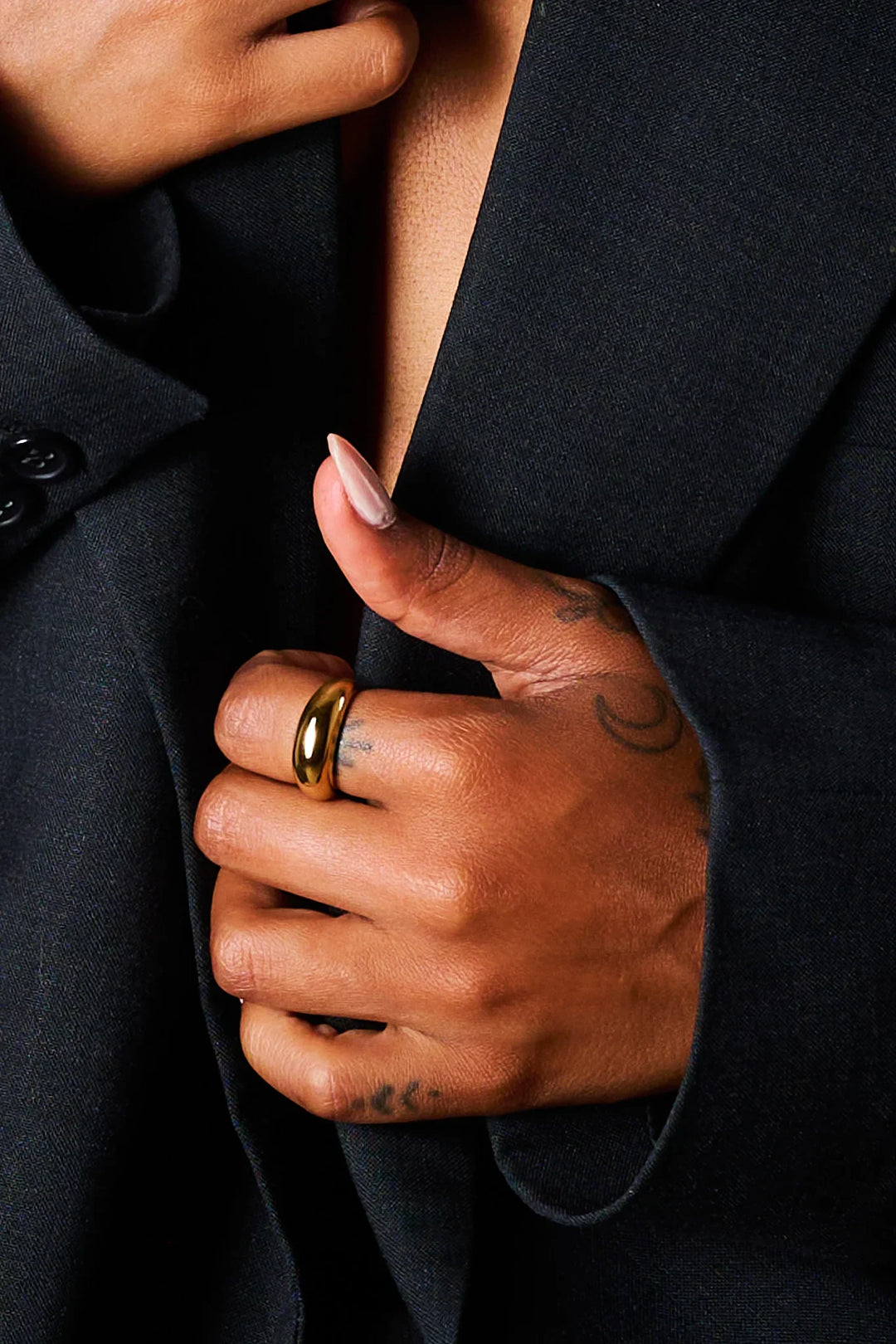 Close-up of hand with a gold dome ring and tattoos, wearing a black blazer