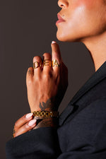 Close-up of hand wearing gold statement rings and bracelet, black blazer, tattoo visible