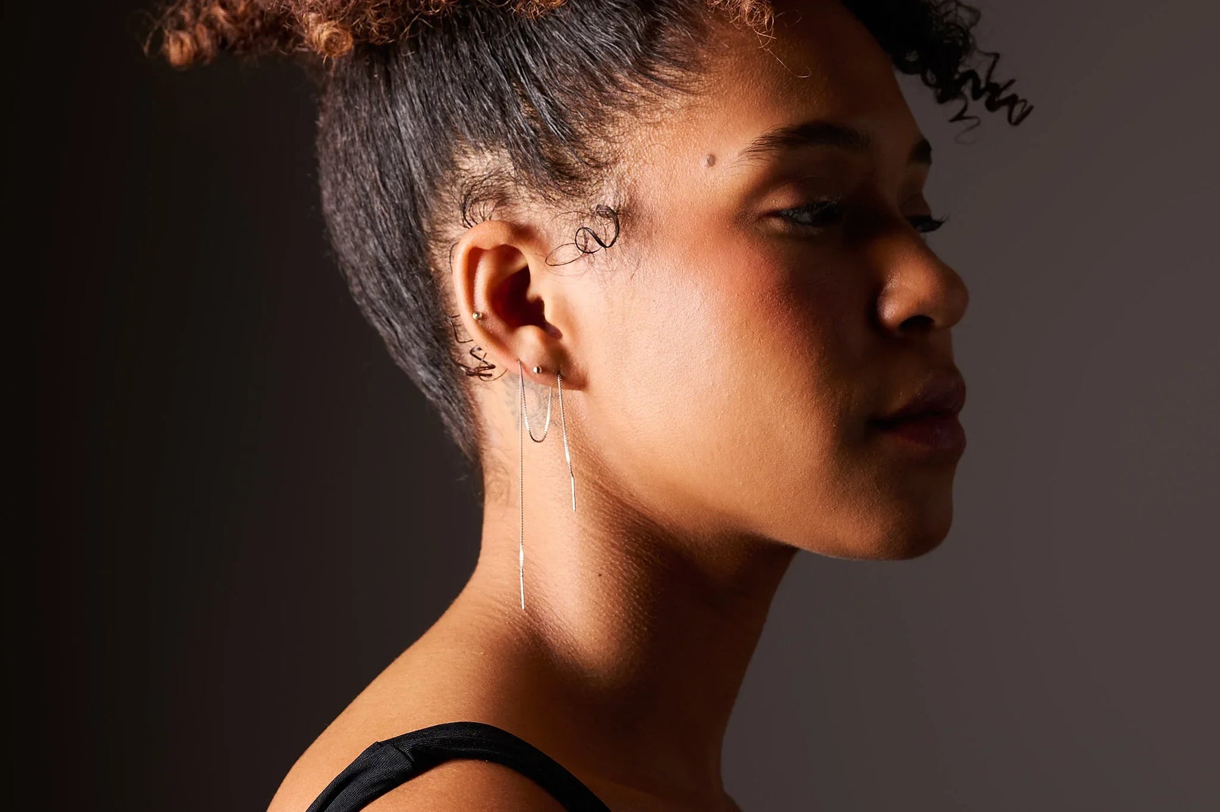 Woman wearing minimalist silver thread earrings and gold ear stud on dark background