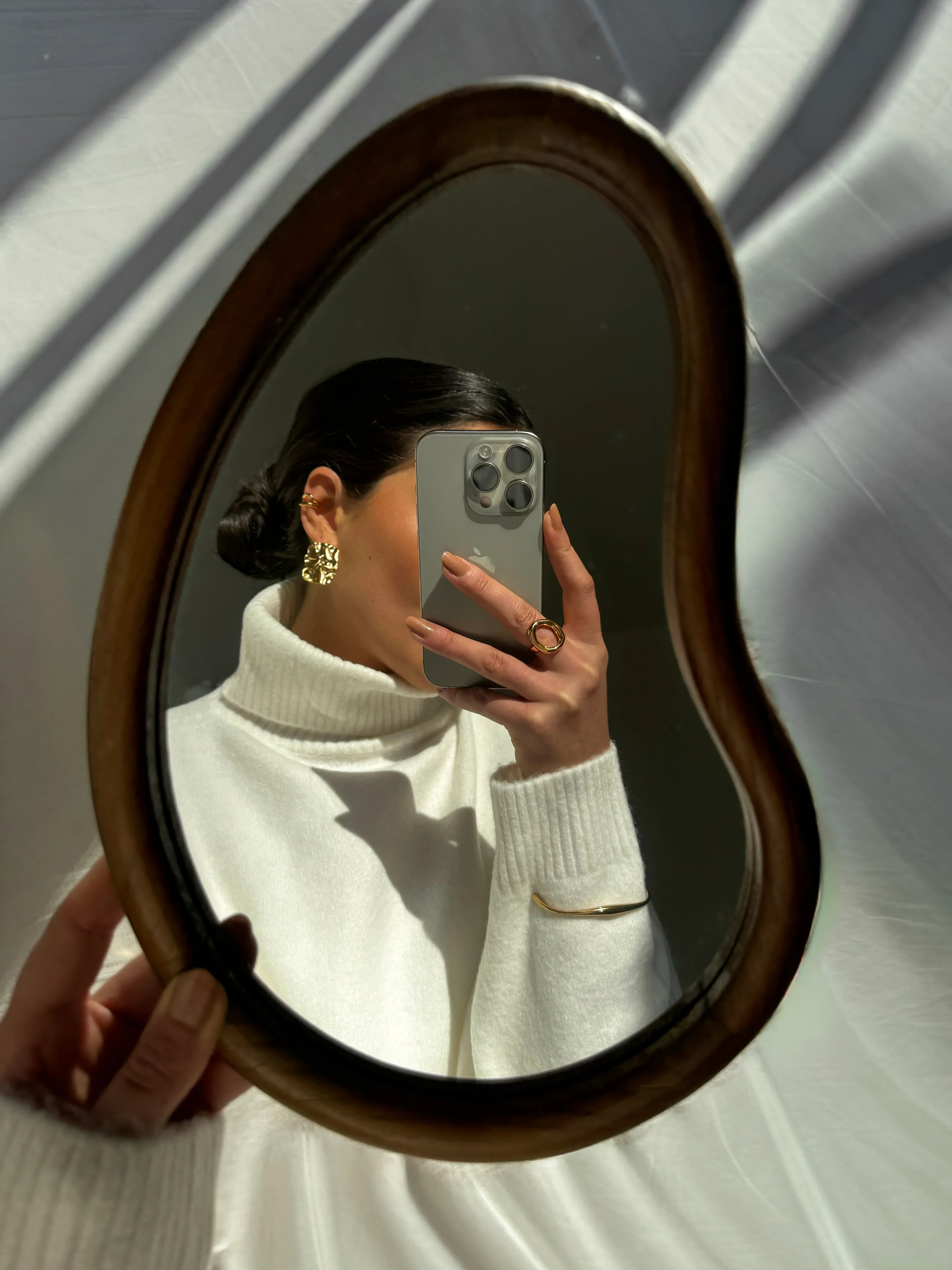 Woman in white turtleneck shows gold earrings, ring, and bracelet in mirror selfie