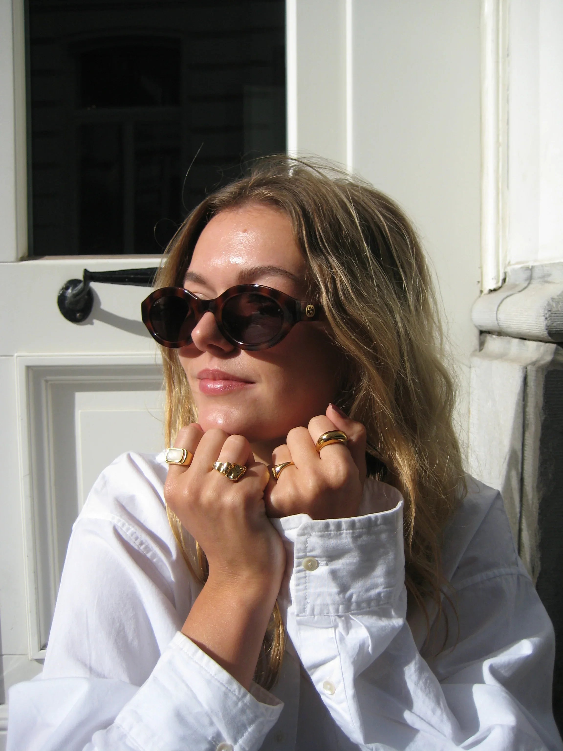 Woman in white shirt wearing sunglasses and gold rings, posing outdoors in sunlight.
