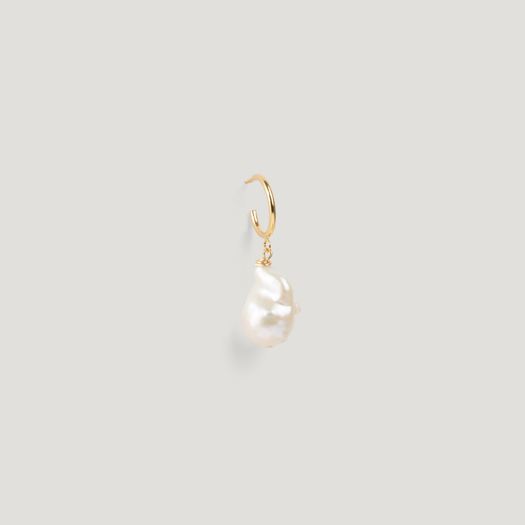 Close-up of a gold hoop earring with an irregular baroque pearl drop on light background.