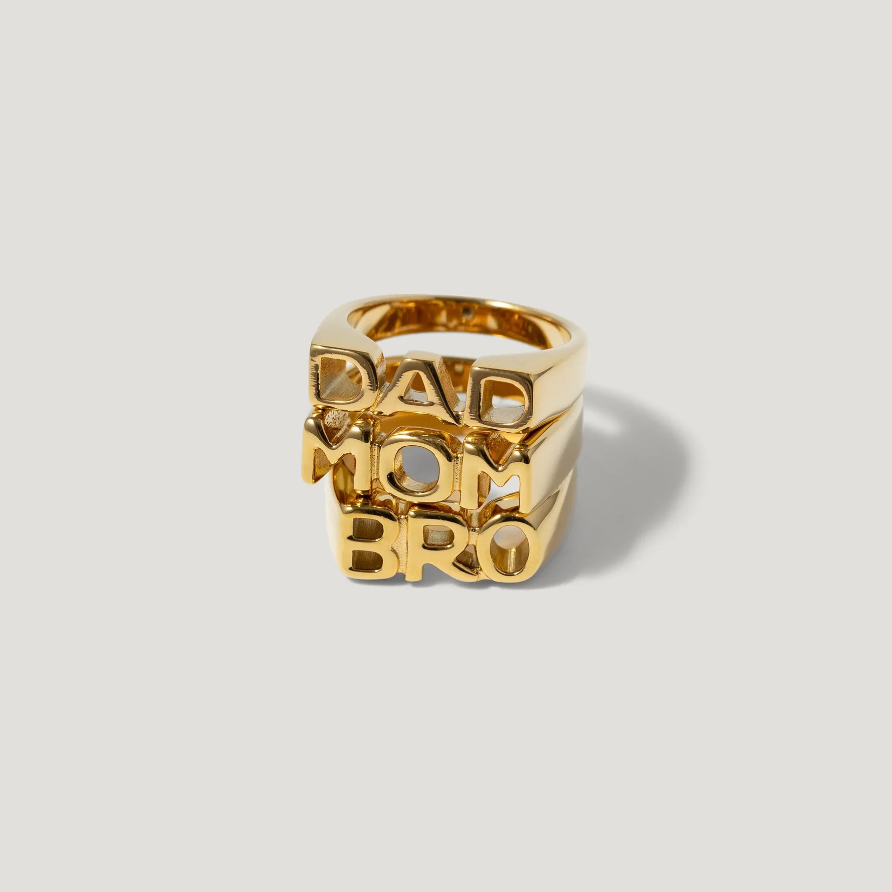 Stacked gold rings with bold DAD MOM BRO letter designs on a light background
