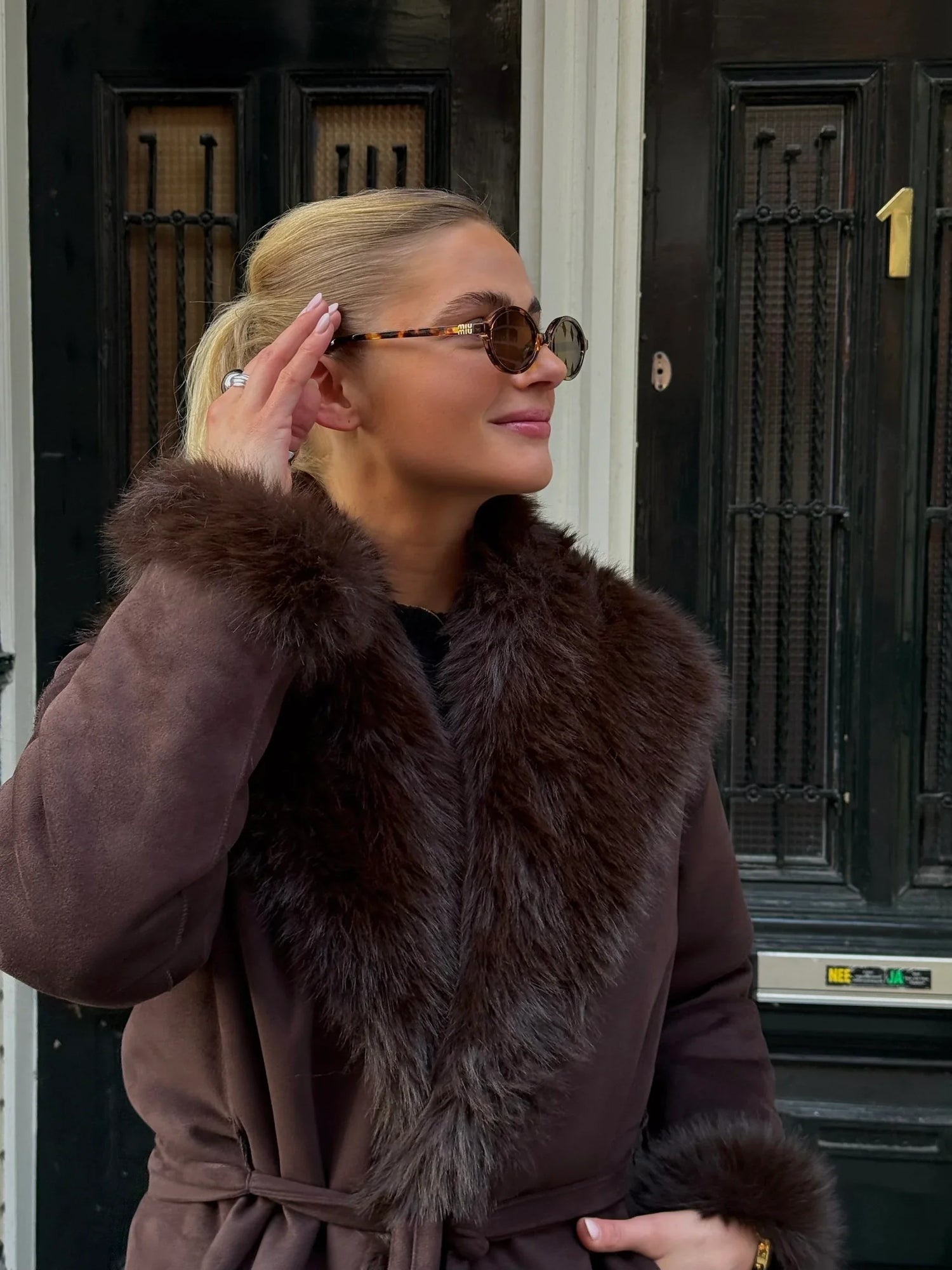 Woman in brown fur-trimmed coat and round sunglasses showcasing silver ring outdoors