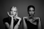 Two women posing in black outfits, showcasing elegant rings, bracelets, and earrings in a studio setting.
