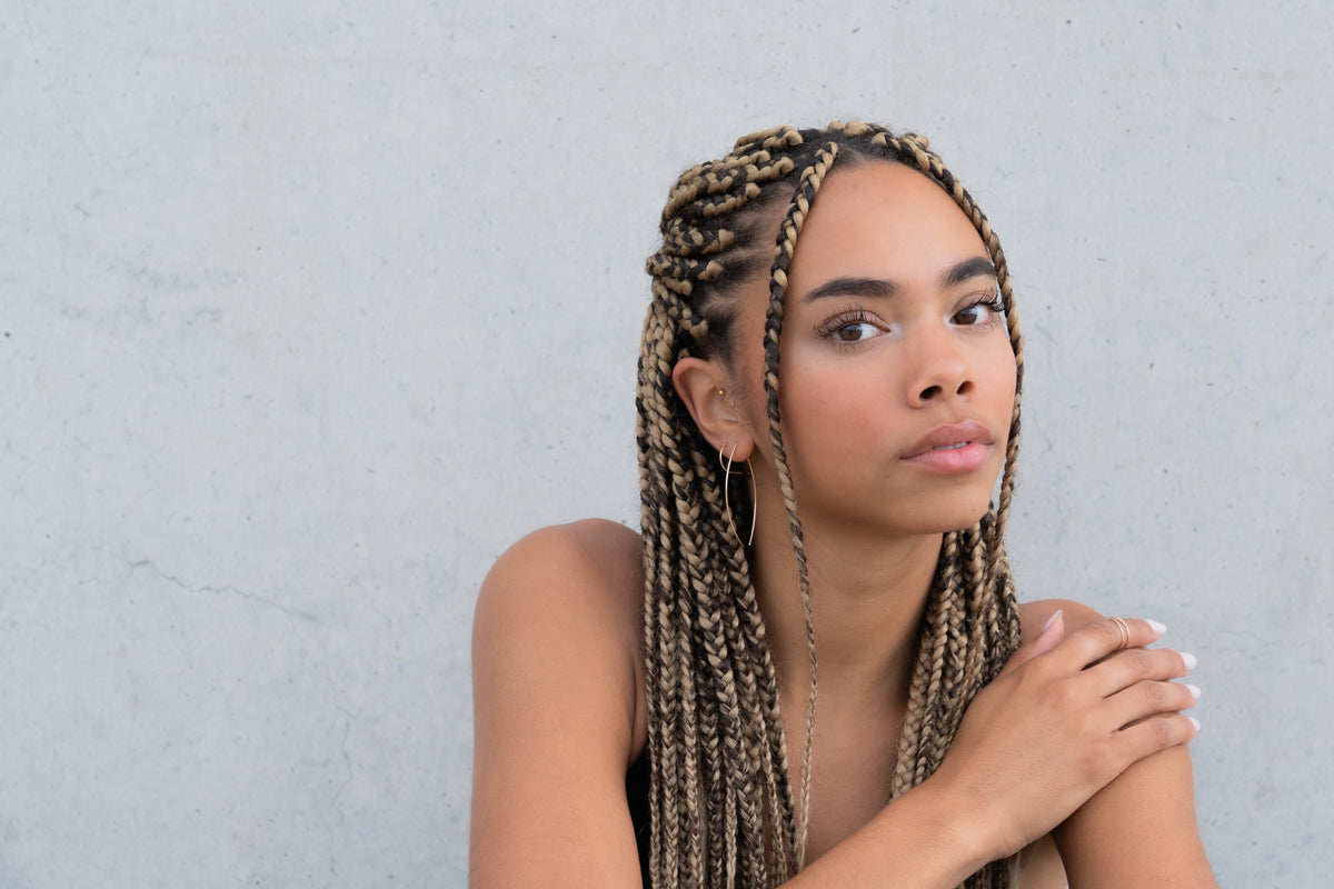 Woman with braided hair wearing a gold thread earring, hand resting on shoulder.