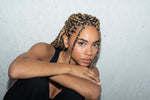 Woman with braided hair wearing a gold thread earring, arms crossed against a wall.