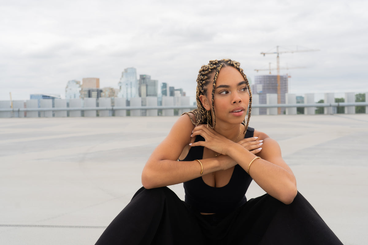 Woman with braided hair crouching, wearing gold bangles on both wrists, city skyline behind.