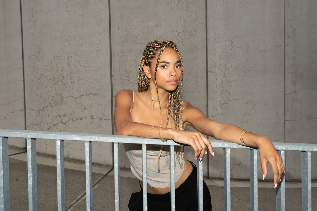 Woman with braided hair leaning on a railing, wearing a gold pearl earring and bracelets.