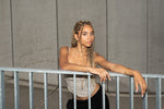 Woman with braided hair leaning on a railing, wearing a gold pearl earring and bracelets.