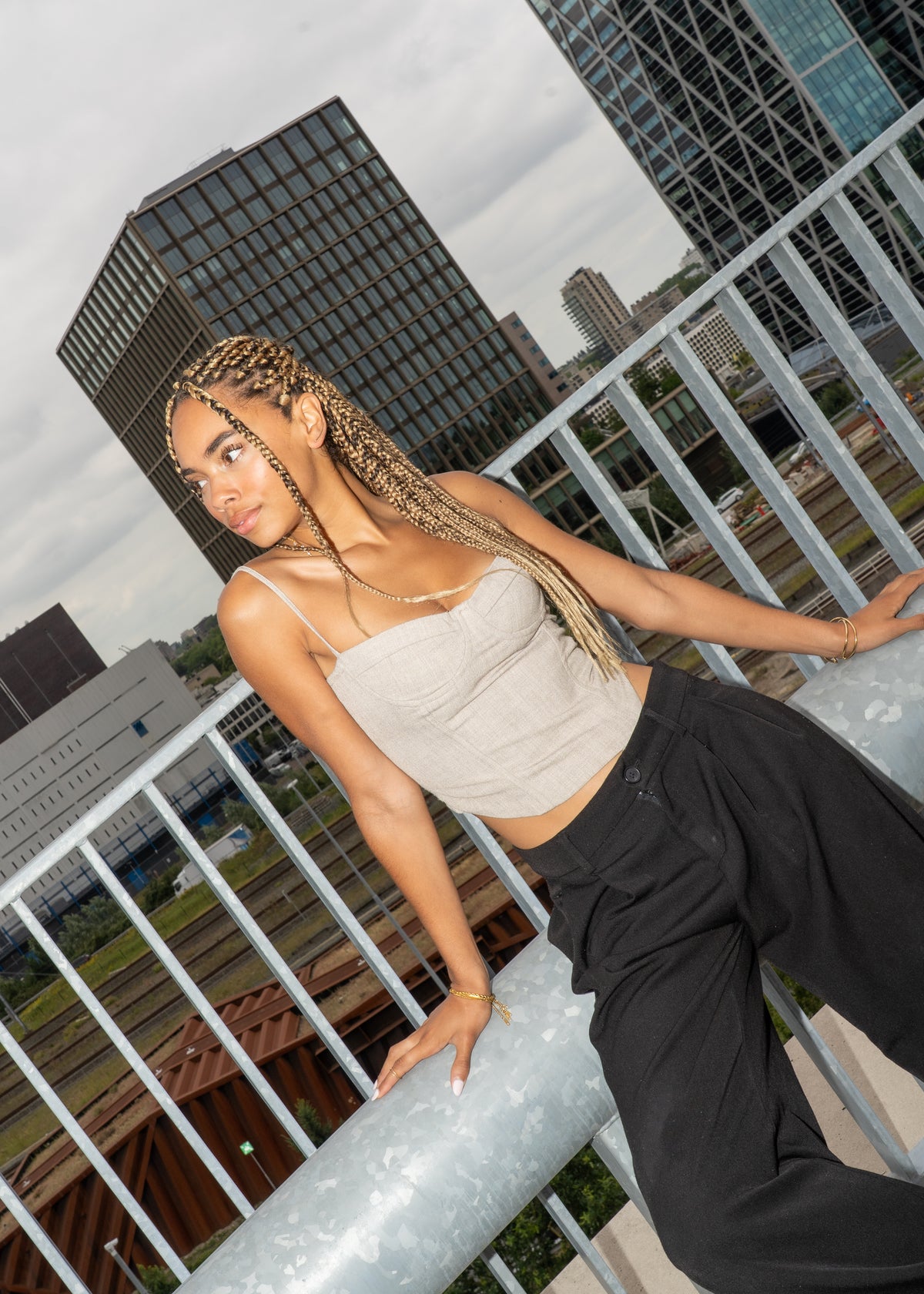 Woman with braided hair leaning on a railing, wearing gold necklace and bracelet, city skyline.