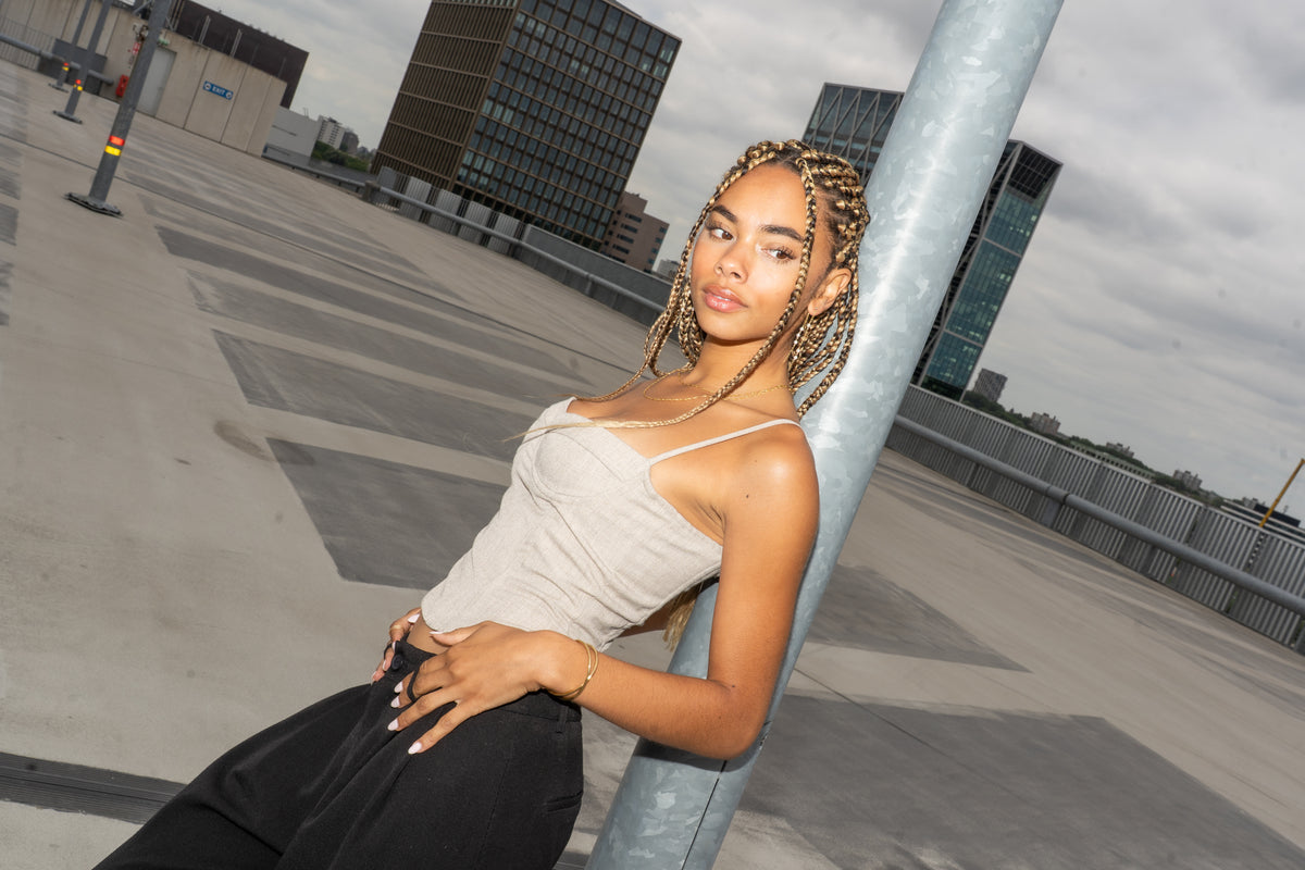 Woman with braided hair leaning against a pole on a rooftop, city skyline behind.