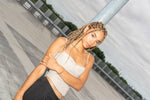 Woman with braided hair, arms crossed, leaning on a rooftop pole, wearing gold pearl earring.