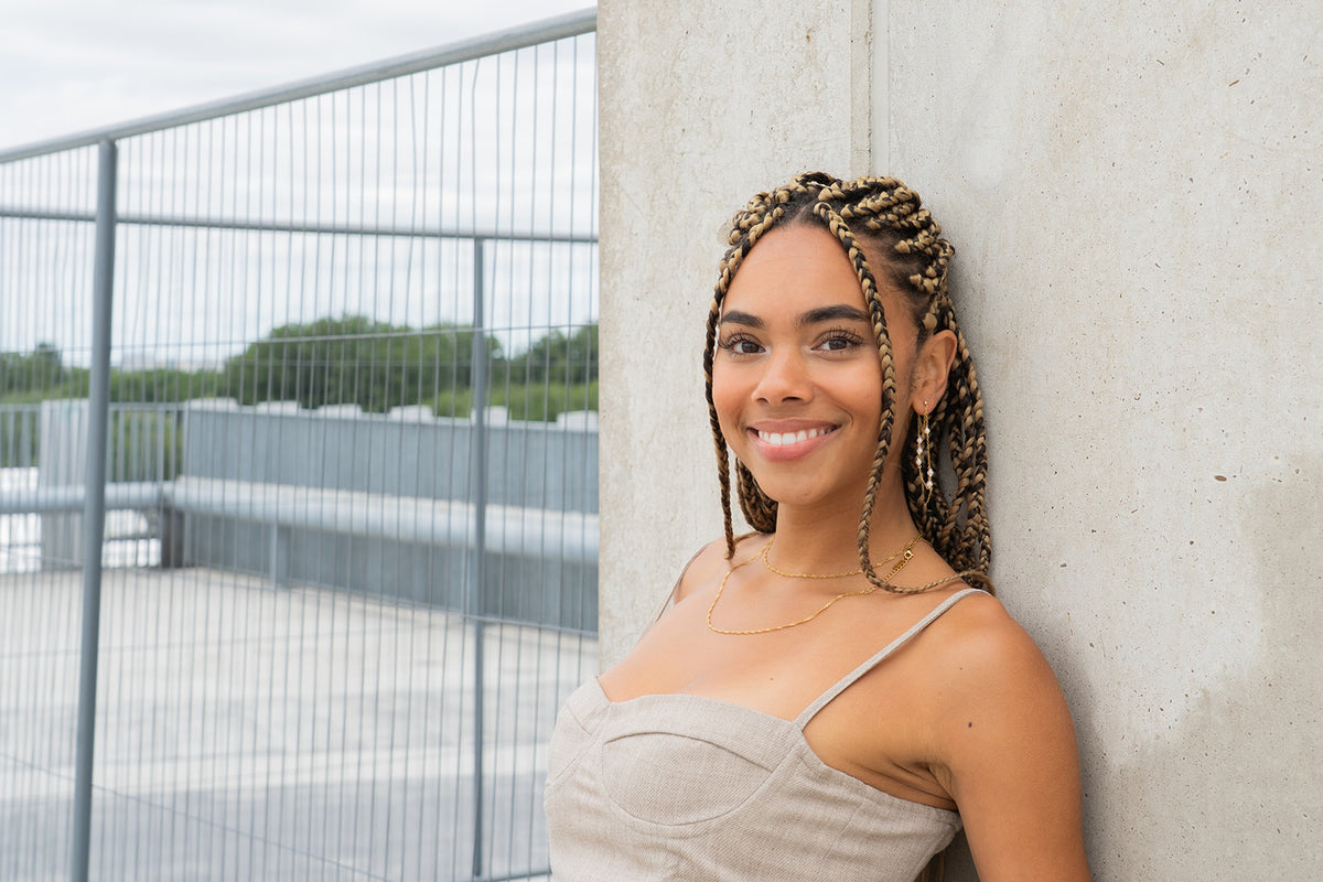 Smiling woman with braided hair wearing a gold pearl earring, leaning against a wall.