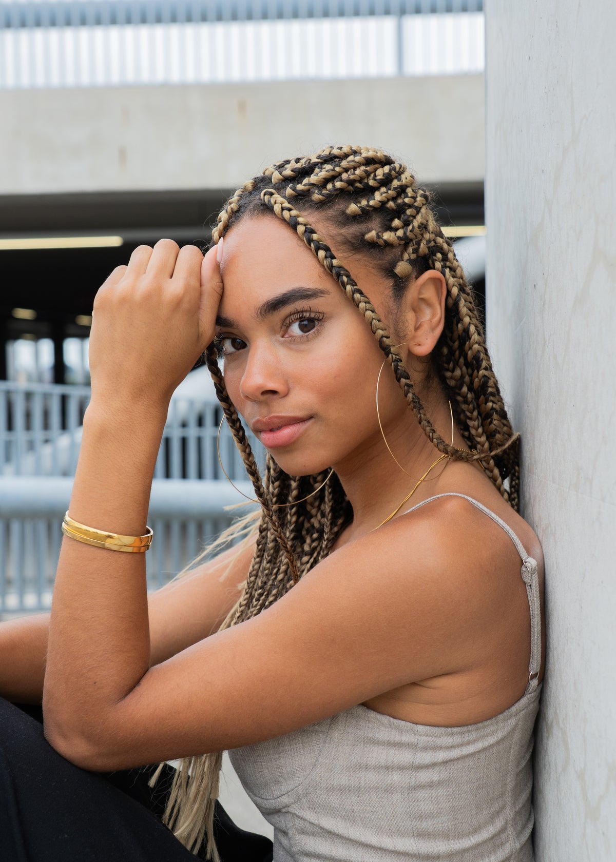 Woman with braided hair wearing a gold hoop earring and bangle, hand on forehead outdoors.