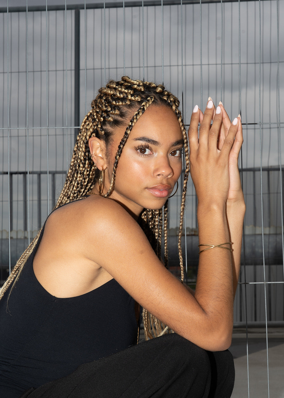 Woman with braided hair wearing a small gold hoop earring and gold bangle, crouching.